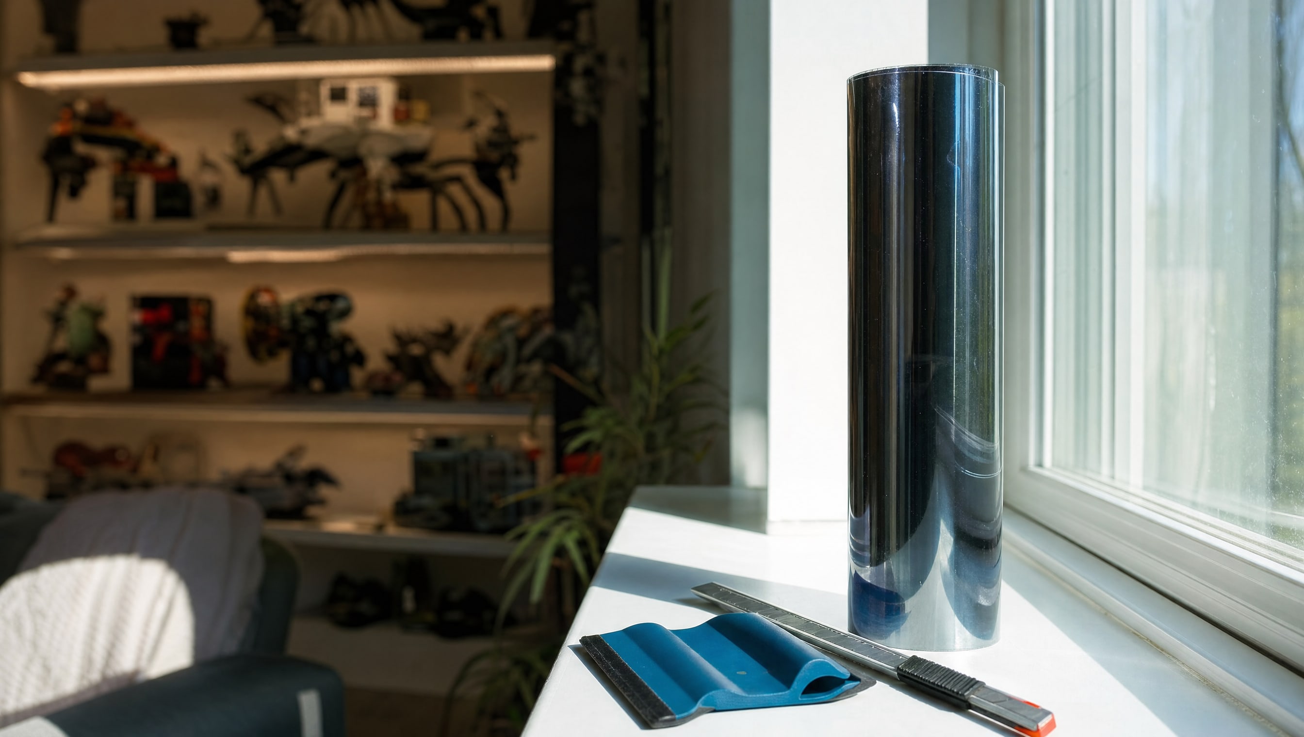 A roll of UV window film stood on a sill beside a bright window, a squeegee and blade alongside.