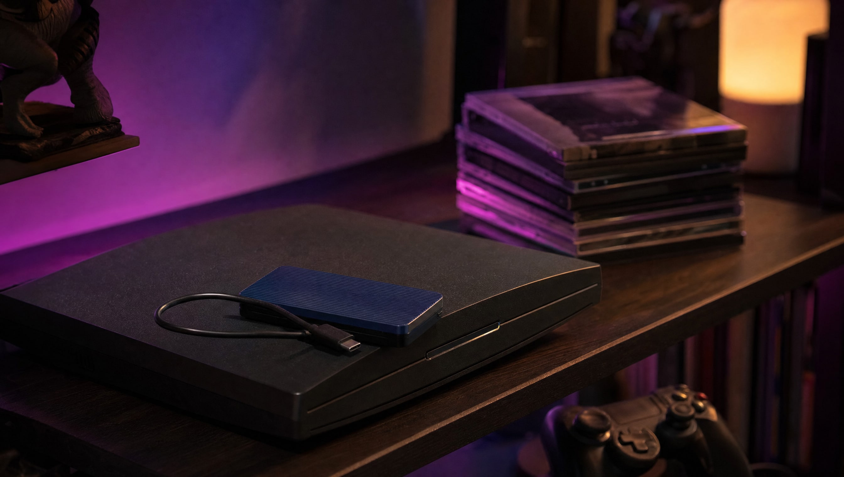 A shelf at night: a flat console, an external drive, a short cable and slim cases.
