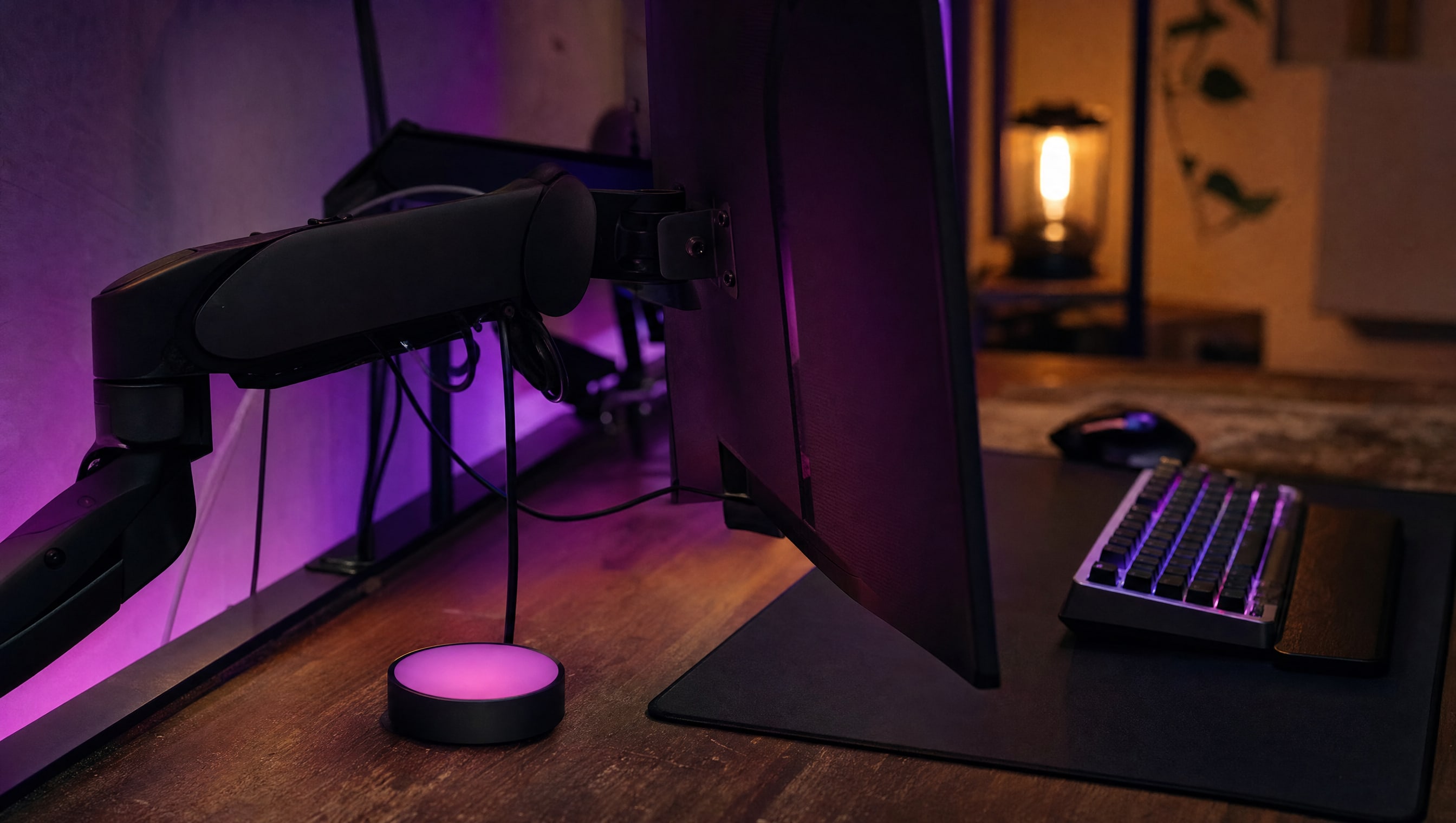 A desk at night: a monitor on an arm, a colour puck on the desk, cables behind.