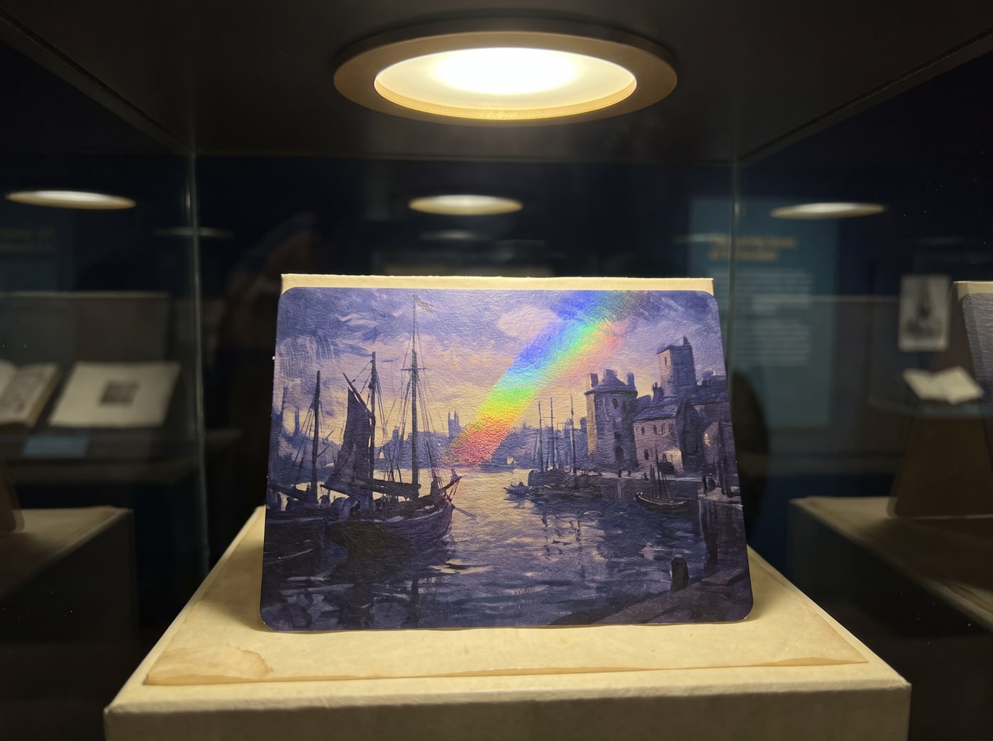 A single card under museum glass in a dark gallery, lit by one warm spot, a painted harbour scene on its face and a rainbow foil shimmer raking across it.