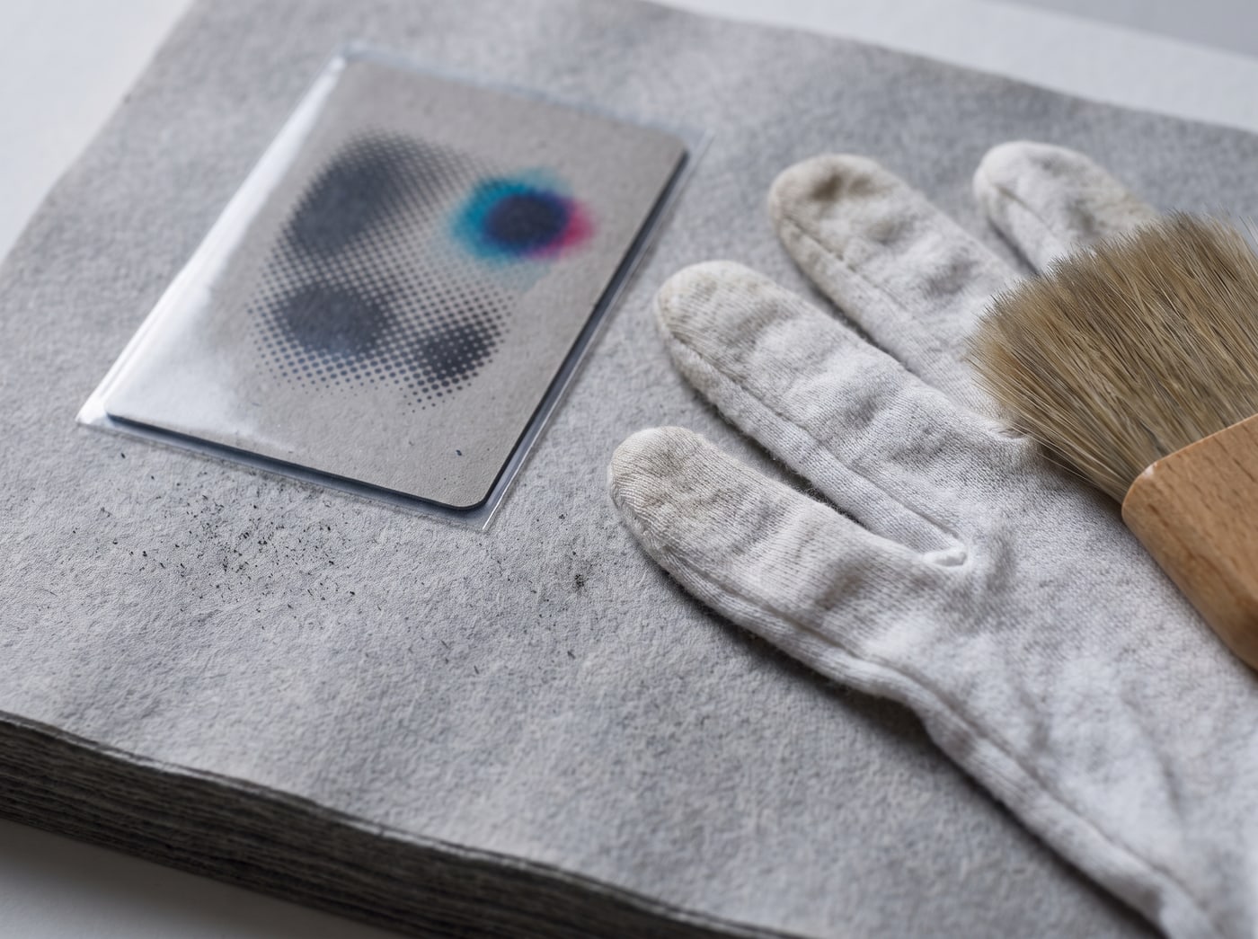 A pair of cotton archivist gloves and a soft goat-hair brush laid on a stack of grey blotting card, beside one sleeved trading card printed in coarse halftone.