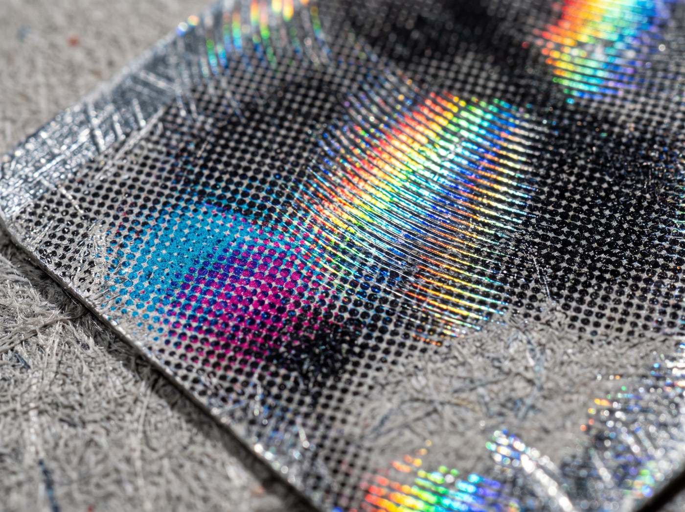 Extreme macro of a foil card surface breaking a lamp into a hard spectrum across a field of printed halftone dots.