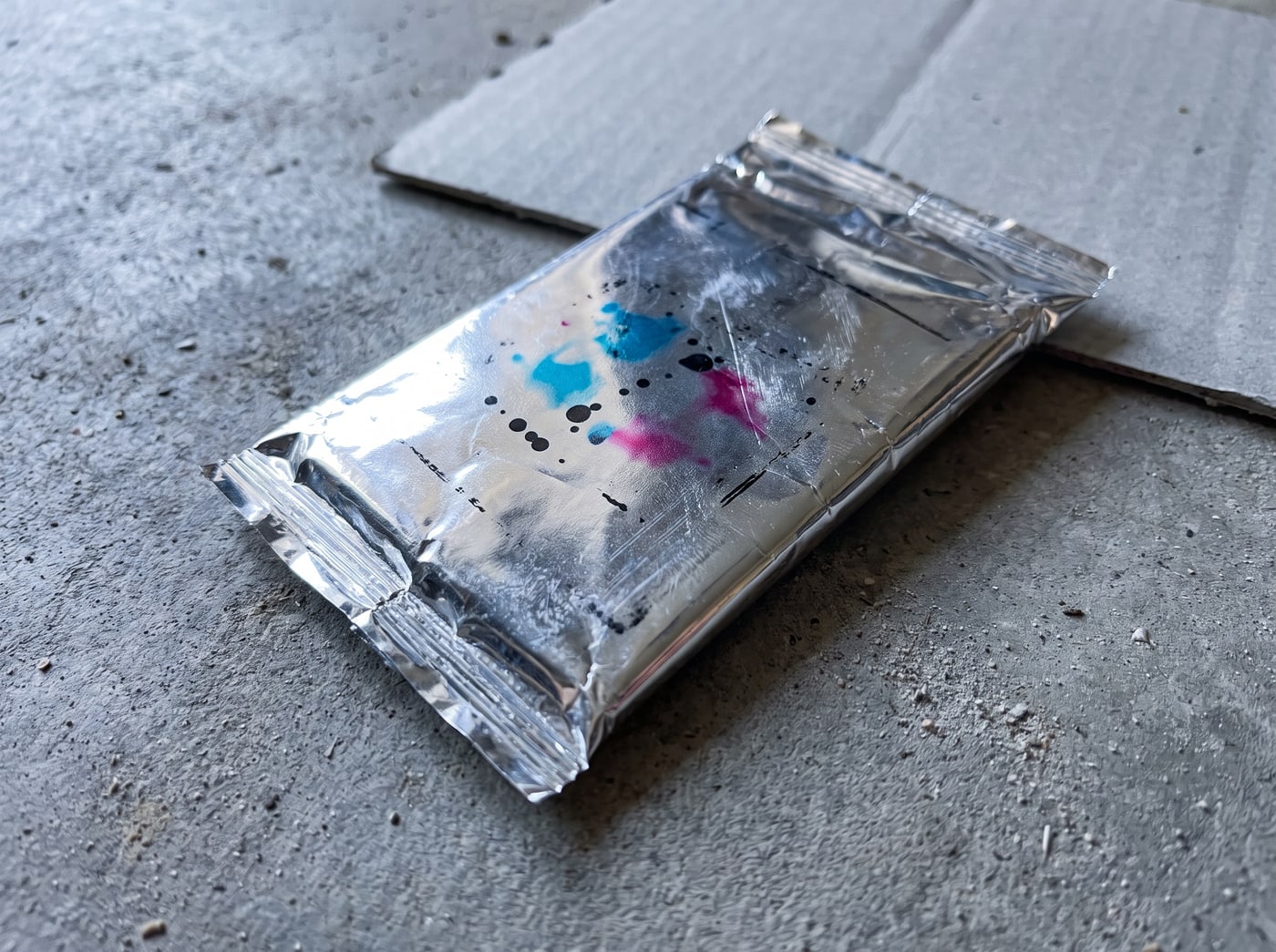 A sealed foil trading card pack lying on grey concrete under a hard flash, the wrapper creased and reflecting.