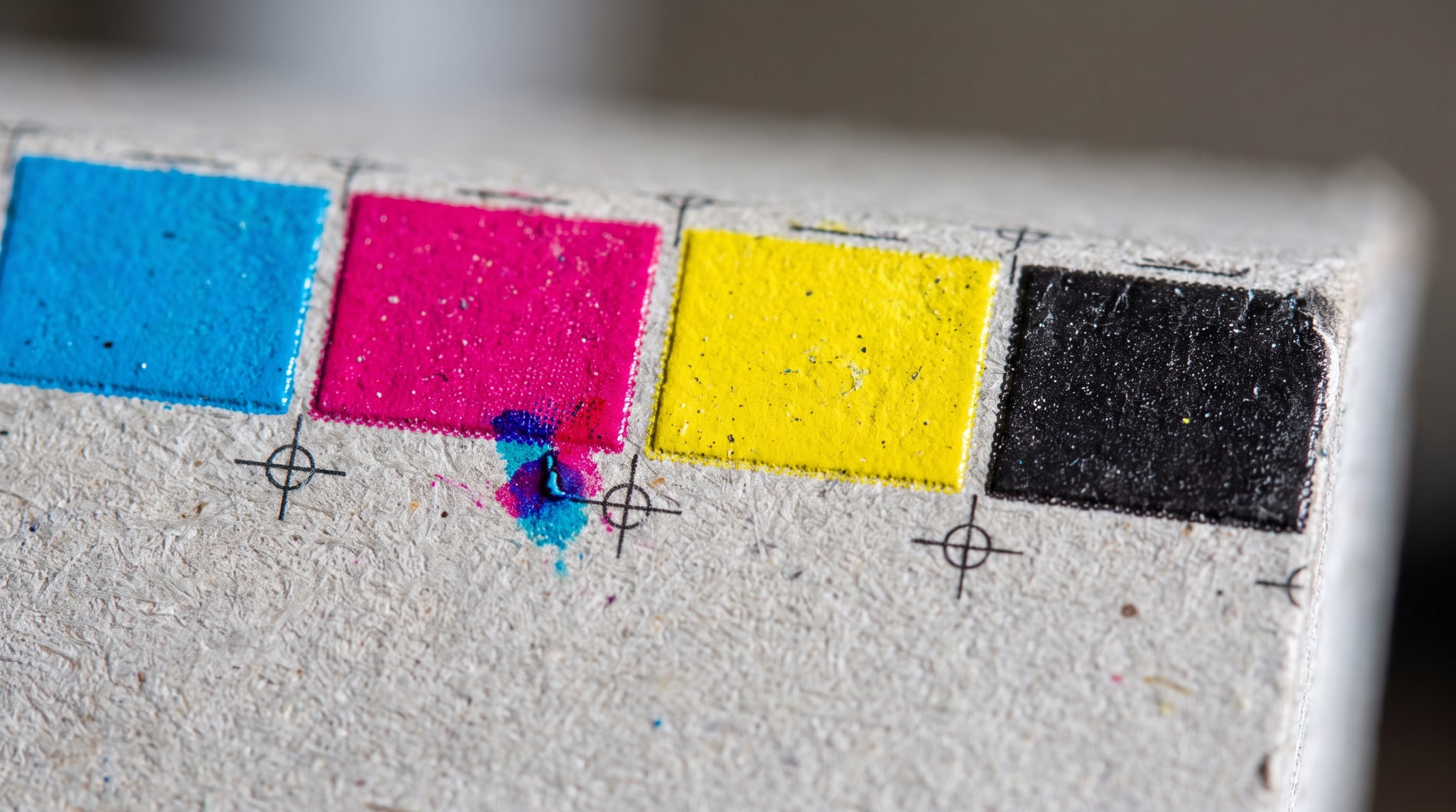 Macro of a printer’s process colour bar on the trimmed edge of grey cardboard: solid blocks of cyan, magenta, yellow and black with registration crosshairs between them.
