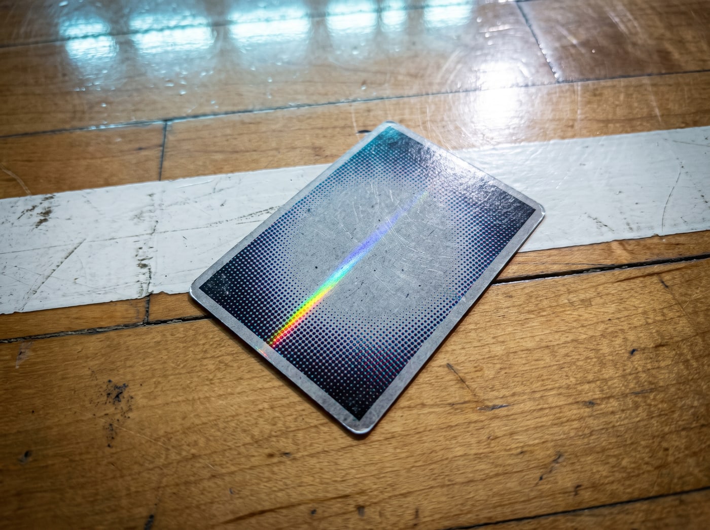 Polished arena boards with a painted white line, hard overhead light, one glossy trading card face down throwing a thin rainbow across the grain.