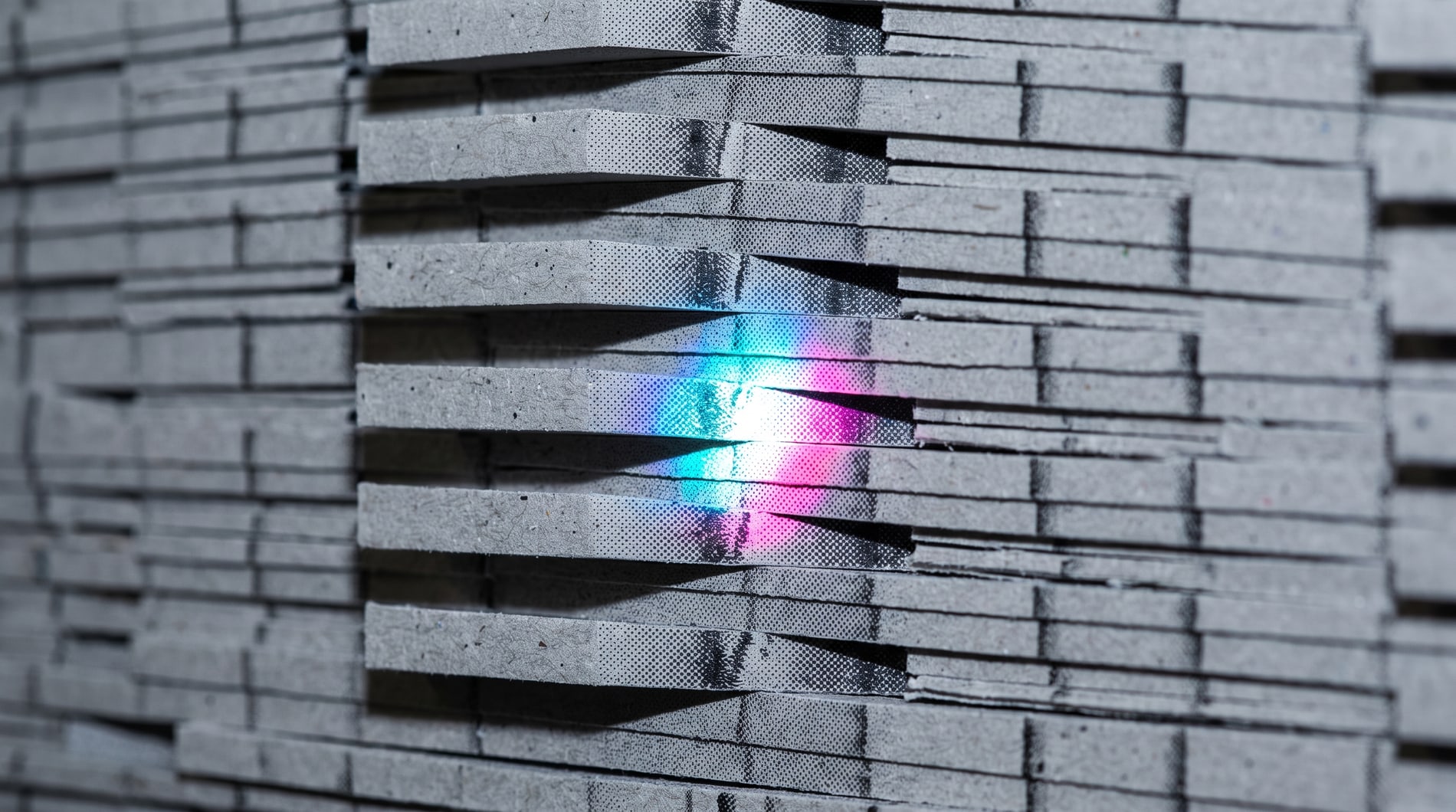 An enormous wall of grey card stock stacked on end, thousands of identical cut edges receding, one narrow strip catching cyan and magenta.