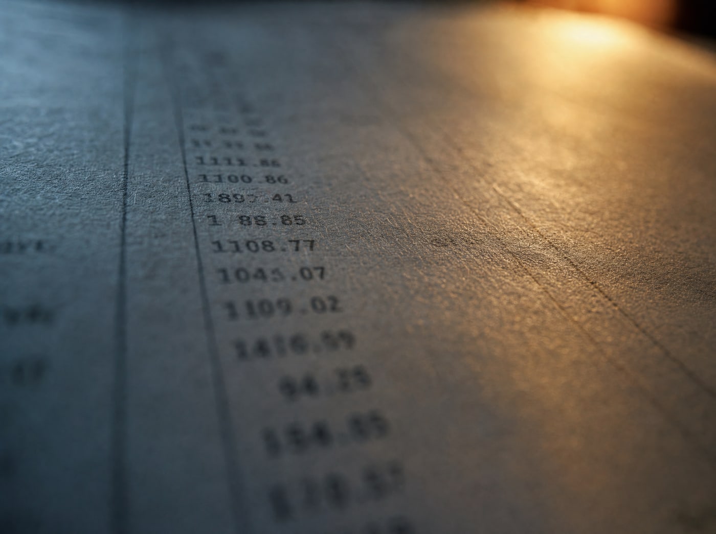 Macro on a printed column of figures on paper, lamplight raking across the fibre, the numbers soft and out of focus.