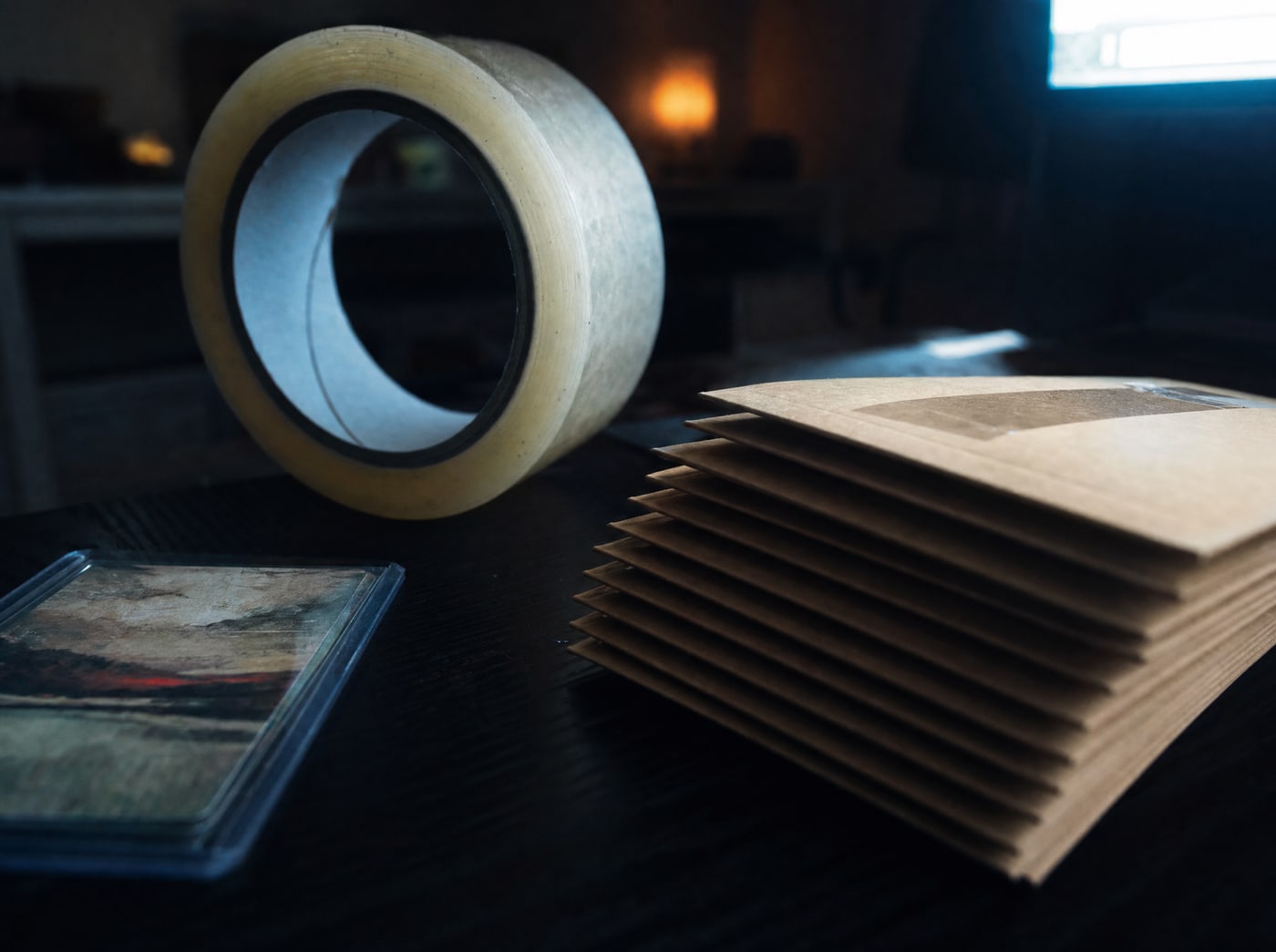 A roll of packing tape standing on end beside a stack of flat rigid mailers under hard side light.