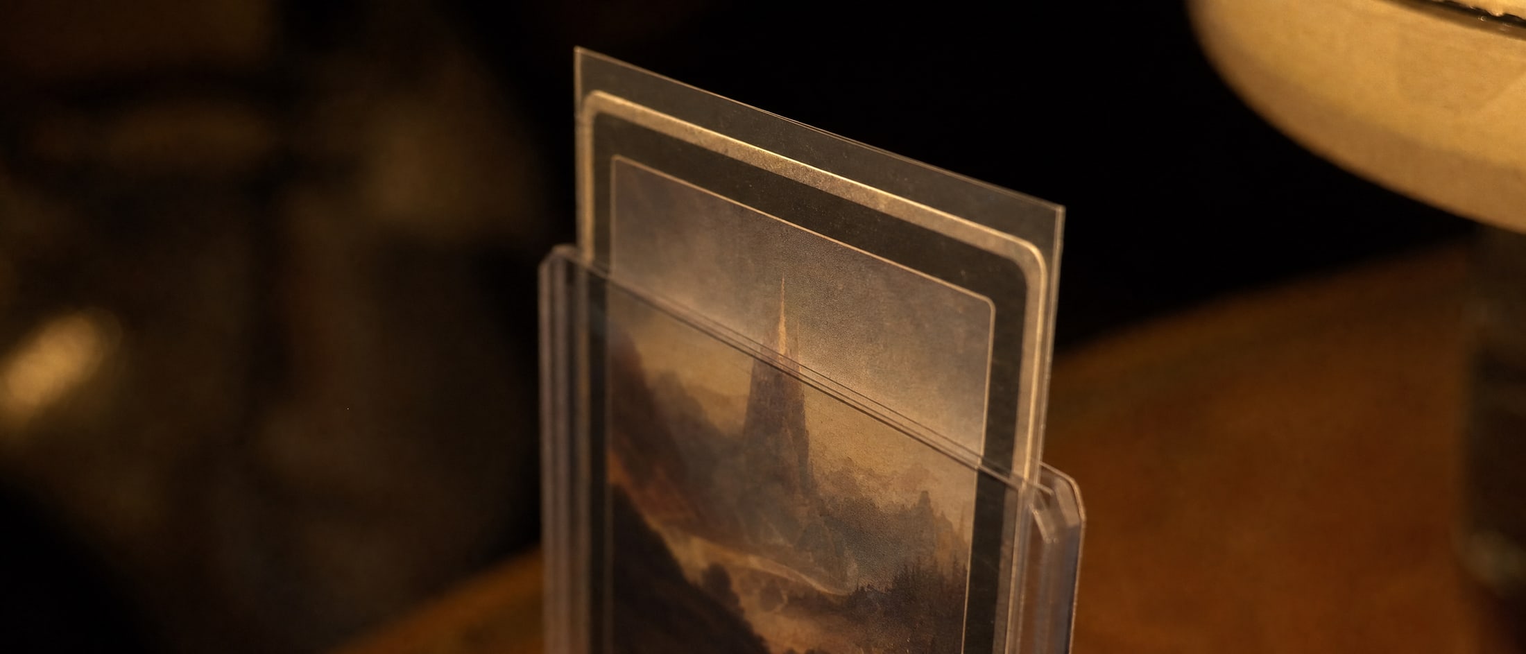 A trading card carrying an invented painted landscape, held in a clear sleeve and slid halfway into a rigid toploader, photographed edge-on under a warm desk lamp so the card, the sleeve and the rigid shell read as three separate layers.